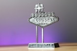 A miniature replica of the iconic 'Welcome to Fabulous Las Vegas, Nevada' sign, featuring detailed lettering and a star on top, set against a soft gradient background transitioning from gray to purple.