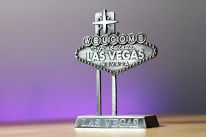 A miniature replica of the iconic 'Welcome to Fabulous Las Vegas, Nevada' sign, featuring detailed lettering and a star on top, set against a soft gradient background transitioning from gray to purple.