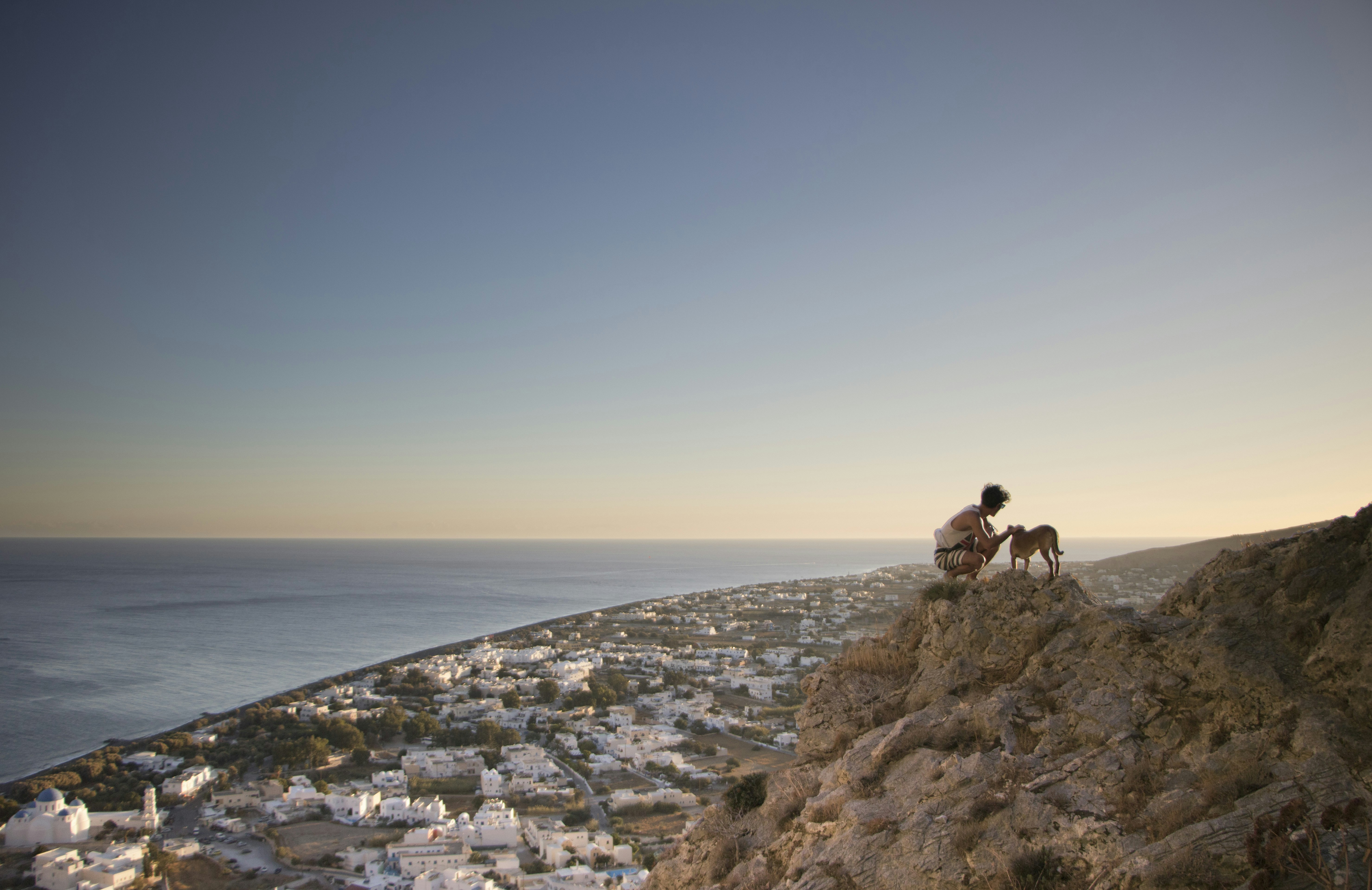 man sitting while holding dog on rock, After a quick look at the local chapel in Santorini, we found a good spot to take a look at the view in the sunset. This picture makes me feel free and one gets a sense of how big the world is, because even from this very tall cliff, we still couldn’t see this tiny island as a whole, just a beach.