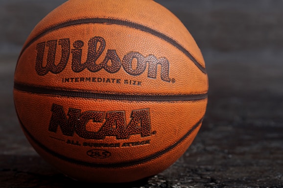 A close-up view of an orange basketball with the brand name Wilson and NCAA logo printed on it. The basketball is positioned against a dark, textured background.
