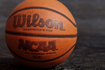 A close-up view of an orange basketball with the brand name Wilson and NCAA logo printed on it. The basketball is positioned against a dark, textured background.