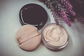 A jar of face powder with a beige makeup puff rests beside a closed, dark-lidded container. Sprigs of purple flowers, possibly lavender, are artfully positioned nearby on a light-colored surface.