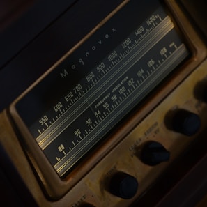 A close-up view of a vintage Magnavox radio tuner featuring a frequency dial with numbers ranging from 550 to 1600. The radio is characterized by a dark brown and gold color scheme, with clearly visible knobs for adjusting settings.