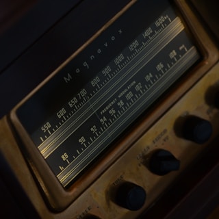 A close-up view of a vintage Magnavox radio tuner featuring a frequency dial with numbers ranging from 550 to 1600. The radio is characterized by a dark brown and gold color scheme, with clearly visible knobs for adjusting settings.