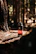 A black water bottle with a red and white logo stands on the forest floor covered in pine needles. Sunlight filters through the trees, creating a warm, natural backdrop. The surrounding trees and greenery are slightly out of focus, enhancing the bottle's prominence.