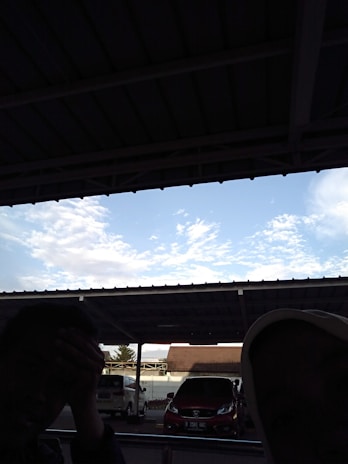 A covered parking area with vehicles visible beneath a large metal roof. The sky is clear with scattered clouds. Part of a person's silhouette is seen on the left, sitting in a shaded area.