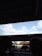 A covered parking area with vehicles visible beneath a large metal roof. The sky is clear with scattered clouds. Part of a person's silhouette is seen on the left, sitting in a shaded area.