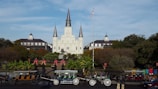 An iconic cathedral with three spires stands prominently in the center, surrounded by greenery and historical buildings. In the foreground, horse-drawn carriages line up along a street, and an American flag flies on a tall pole.