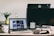 A workspace features a laptop displaying a gallery of images, a desktop monitor, a tablet with text, and various objects like a plant, water bottle, mug, two small figurines, a smartphone, and a computer mouse. The setting is well-lit with natural light.