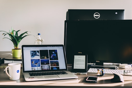 A workspace features a laptop displaying a gallery of images, a desktop monitor, a tablet with text, and various objects like a plant, water bottle, mug, two small figurines, a smartphone, and a computer mouse. The setting is well-lit with natural light.