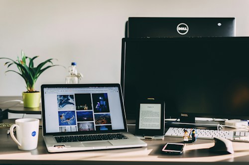 A workspace features a laptop displaying a gallery of images, a desktop monitor, a tablet with text, and various objects like a plant, water bottle, mug, two small figurines, a smartphone, and a computer mouse. The setting is well-lit with natural light.