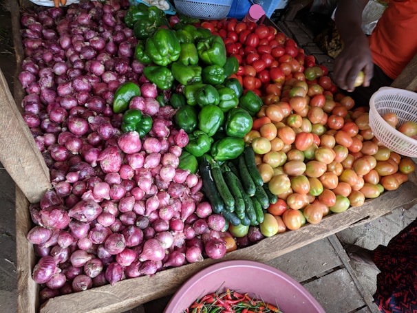 A wooden stall displaying an assortment of vegetables including red onions, green bell peppers, cucumbers, tomatoes in shades of green and red, and a basket filled with red chili peppers. A person's hand is seen picking a tomato, adding a human element to the bustling market scene.