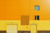 A bright orange and yellow building facade with two doors and two windows. The structure has a clean, geometric design, featuring a grid-like pattern of panels. One window is positioned above each door, both with a blue-tinted glass. The vibrant colors create a striking contrast with the snow visible at the base of the building.