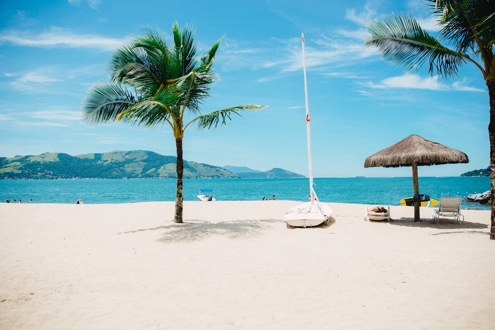 A leaning palm, a small sailing boat and a thatched umbrella on white sand by turquoise water