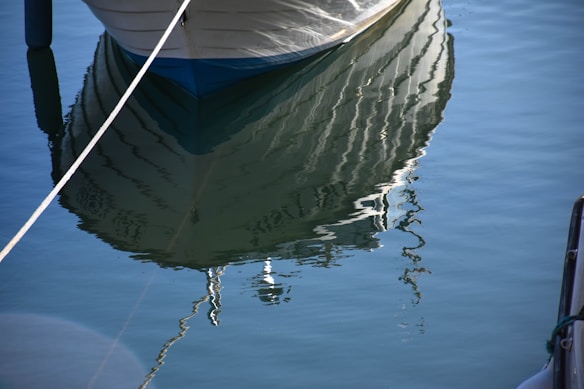 The image depicts the reflection of a boat's hull in calm water, creating a mirror-like effect with slight ripples. A rope is visible, securing the boat to a dock. The water is clear, allowing the reflection to appear with some distortion.
