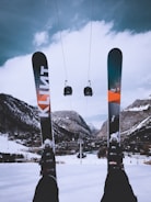 A snowy mountain landscape with two skis in the foreground and a pair of gondolas suspended on a cable in the midground. A village sits nestled in the valley surrounded by snow-covered mountains under a cloudy sky.