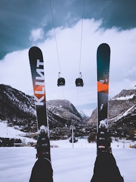 A snowy mountain landscape with two skis in the foreground and a pair of gondolas suspended on a cable in the midground. A village sits nestled in the valley surrounded by snow-covered mountains under a cloudy sky.