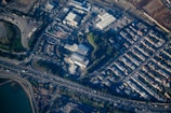 An aerial view of an urban area featuring a cluster of industrial buildings, a highway with moving vehicles, and a densely packed residential neighborhood. There are patches of greenery interspersed among the structures, and water is visible at the bottom edge.