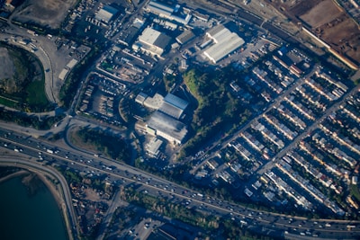 An aerial view of an urban area featuring a cluster of industrial buildings, a highway with moving vehicles, and a densely packed residential neighborhood. There are patches of greenery interspersed among the structures, and water is visible at the bottom edge.