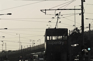 A silhouette of a tram with overhead electrical wires against a cloudy sky. The foreground features multiple intersecting cables and poles, creating a complex network above the city street. In the background, an overpass and various street lamps add to the urban landscape.