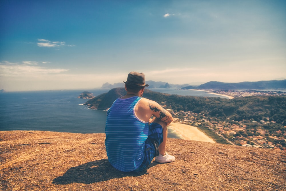 man sitting on hill facing body of water under clear sky