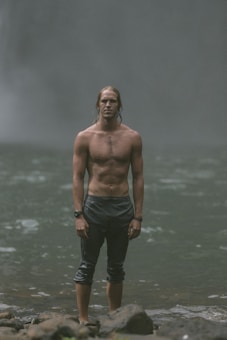 A shirtless man stands confidently in a rocky, misty setting. His hair is wet, and he is wearing rolled-up dark pants and a watch on his wrist. The background suggests a waterfall or forested area with a muted, foggy atmosphere.