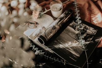 An artistic arrangement featuring an open book with handwritten notes, a pair of eyeglasses, a lit candle, a flower, and a black-and-white photograph. The scene is further adorned with delicate leaves and a warm, soft backdrop.