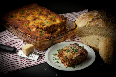 A freshly baked lasagna in a glass dish, topped with herbs, sits on a red and white checkered cloth. Beside it, a white plate holds a serving of lasagna, garnished with fresh parsley and grated cheese. A loaf of crusty bread is partially sliced, and a block of cheese rests on a grater with a black handle.