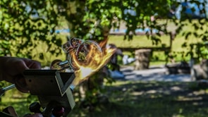 A detailed image capturing a moment in the glassblowing process. Hands are manipulating a glowing piece of molten glass, which is being shaped with the assistance of a tool. The background features a lush, green, outdoor setting with dappled sunlight filtering through the trees, and blurred elements indicating a bench and other items in the distance.