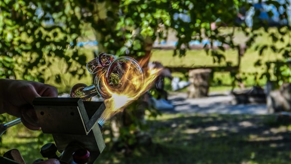 A detailed image capturing a moment in the glassblowing process. Hands are manipulating a glowing piece of molten glass, which is being shaped with the assistance of a tool. The background features a lush, green, outdoor setting with dappled sunlight filtering through the trees, and blurred elements indicating a bench and other items in the distance.