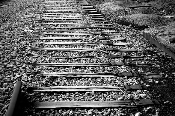 Wooden railroad tracks stretch into the distance, bordered by gravel and natural terrain. The image is in black and white, highlighting the texture of the wooden sleepers and the surrounding stones.