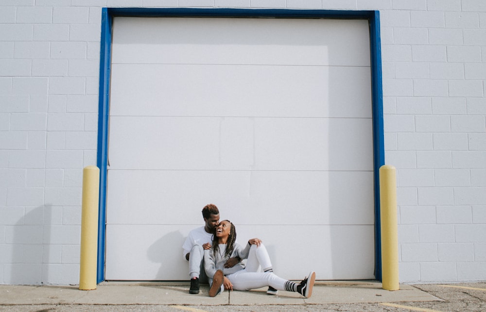 man and woman sitting on ground while leaning on white wall