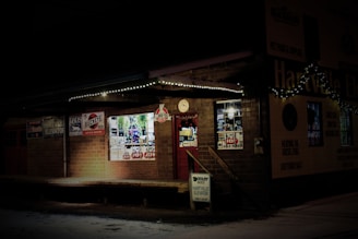A dimly-lit storefront with brick walls decorated for the holiday season. Small white lights are strung along the roof, and a wreath can be seen hanging near the entrance. Various signs and advertisements for products like Buckeye feeds and Dad's creams are displayed on the walls. The windows reveal a glimpse of decorations and merchandise inside.