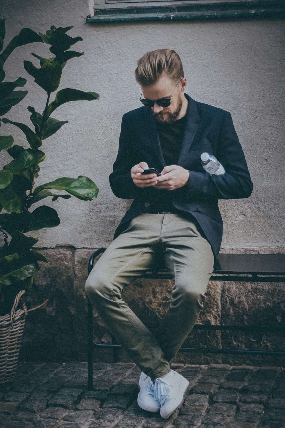 man using smartphone sitting on black metal bench near brown concrete wall