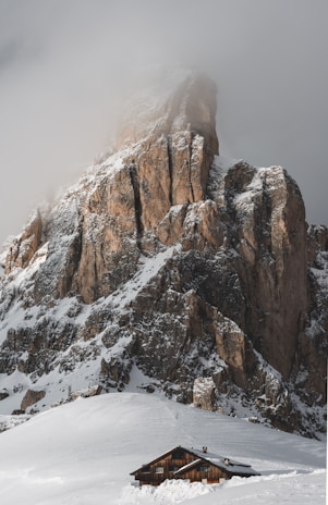 A solitary wooden cabin is nestled at the base of a towering, snow-covered mountain peak. The rugged rock face is partially shrouded in mist, creating a dramatic and serene alpine scene.