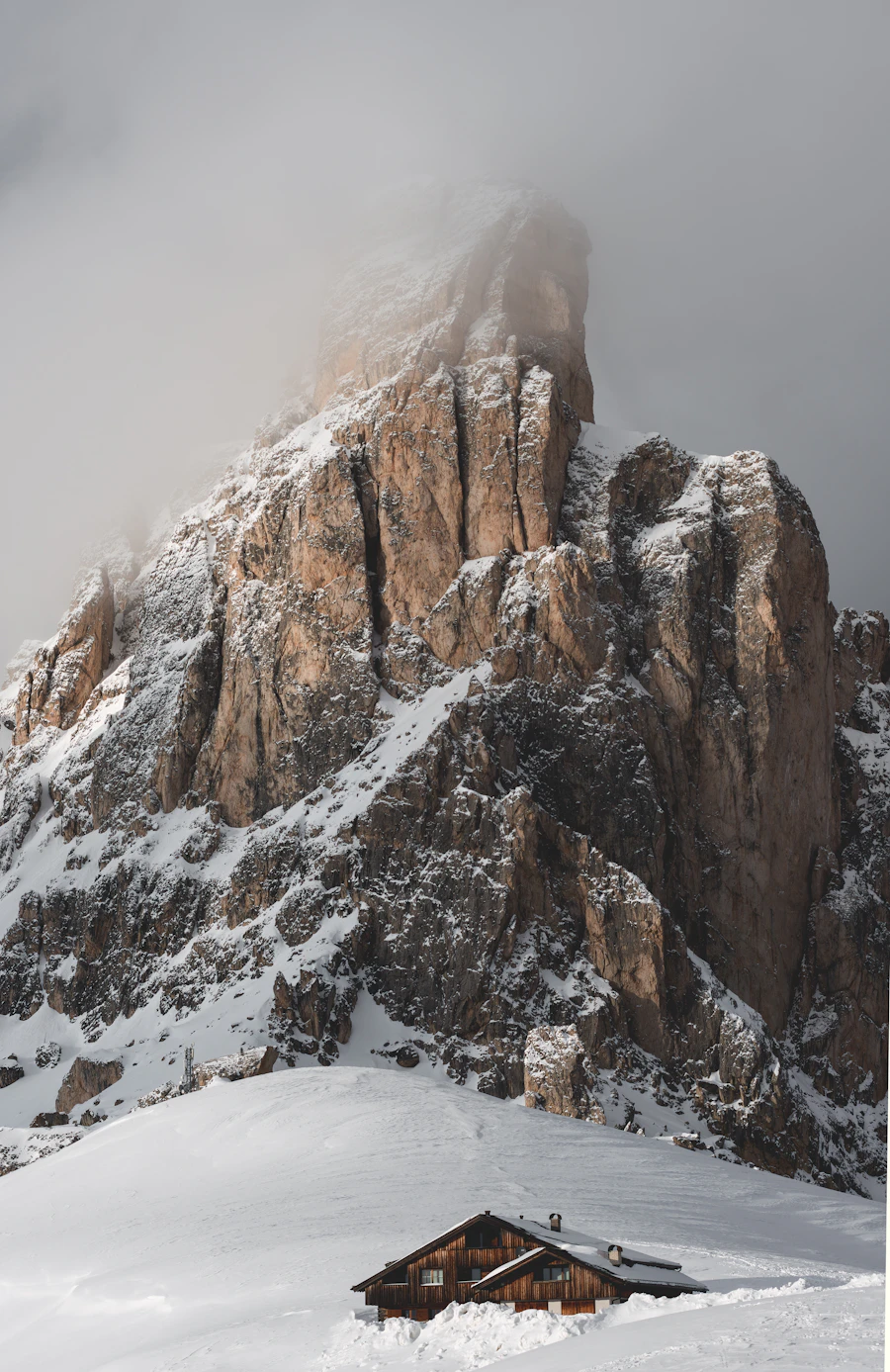 Wooden mountain hut in deep snow below a mist-shrouded Dolomite rock tower