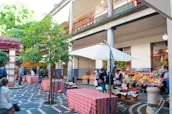 An outdoor marketplace with various fruit and vegetable stalls is bustling with activity. People are browsing and purchasing items under colorful umbrellas and a mix of modern and traditional architectural elements are visible. A large tree and greenery enhance the inviting atmosphere.