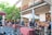 An outdoor marketplace with various fruit and vegetable stalls is bustling with activity. People are browsing and purchasing items under colorful umbrellas and a mix of modern and traditional architectural elements are visible. A large tree and greenery enhance the inviting atmosphere.