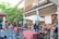 An outdoor marketplace with various fruit and vegetable stalls is bustling with activity. People are browsing and purchasing items under colorful umbrellas and a mix of modern and traditional architectural elements are visible. A large tree and greenery enhance the inviting atmosphere.