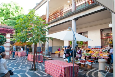 An outdoor marketplace with various fruit and vegetable stalls is bustling with activity. People are browsing and purchasing items under colorful umbrellas and a mix of modern and traditional architectural elements are visible. A large tree and greenery enhance the inviting atmosphere.