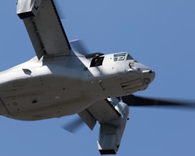 A military aircraft is captured in flight, prominently featuring its tilting rotors and robust design. The sky provides a clear blue backdrop, emphasizing the aircraft's metallic exterior and the motion of the rotors.