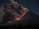 A towering volcano is actively erupting with a massive plume of dark smoke and ash billowing into the sky. Bright orange and red lava flows down the slope, illuminating the otherwise dark landscape. Sparse vegetation is visible in the foreground under a dim, night sky.