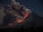 A towering volcano is actively erupting with a massive plume of dark smoke and ash billowing into the sky. Bright orange and red lava flows down the slope, illuminating the otherwise dark landscape. Sparse vegetation is visible in the foreground under a dim, night sky.
