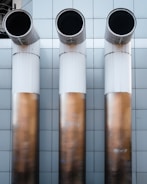 Three large, cylindrical industrial pipes protrude horizontally from a metallic tiled wall. The pipes are positioned side by side and have a clean, modern appearance with a gradient from white to a bronze-like hue.