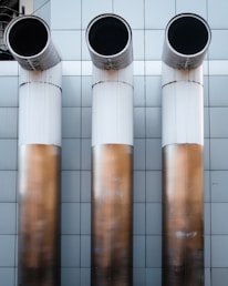 Three large, cylindrical industrial pipes protrude horizontally from a metallic tiled wall. The pipes are positioned side by side and have a clean, modern appearance with a gradient from white to a bronze-like hue.