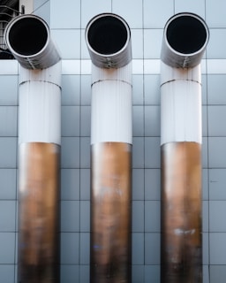 Three large, cylindrical industrial pipes protrude horizontally from a metallic tiled wall. The pipes are positioned side by side and have a clean, modern appearance with a gradient from white to a bronze-like hue.