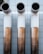 Three large, cylindrical industrial pipes protrude horizontally from a metallic tiled wall. The pipes are positioned side by side and have a clean, modern appearance with a gradient from white to a bronze-like hue.