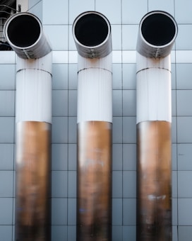 Three large, cylindrical industrial pipes protrude horizontally from a metallic tiled wall. The pipes are positioned side by side and have a clean, modern appearance with a gradient from white to a bronze-like hue.