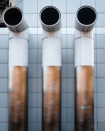 Three large, cylindrical industrial pipes protrude horizontally from a metallic tiled wall. The pipes are positioned side by side and have a clean, modern appearance with a gradient from white to a bronze-like hue.