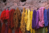 Various strands of brightly colored yarn or threads hang against a textured, earthy wall. The colors range from deep reds and browns to yellows, purples, pinks, and blues. The yarns are arranged in a way that contrasts with the rustic appearance of the background, suggesting a traditional or handmade context.